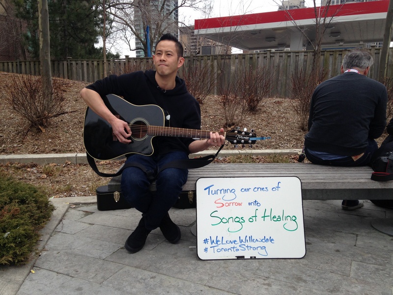 A man with a guitar at the Toronto Willowdale terrorist site. A sign reads, "Turning our cries of sorrow into songs of healing."