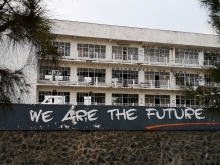 Graffiti, &quot;We are the future&quot; is spray painted on the wall in front of the University of Goma. In the background, half of the windows in the five-story building are broken.